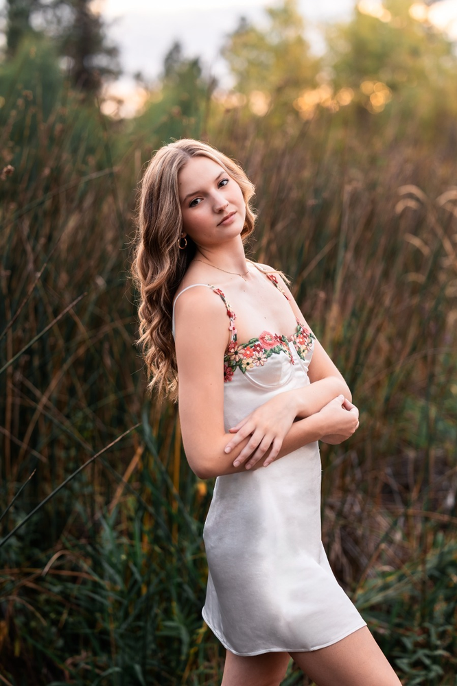 Girl standing with arms crossed wearing white dress in tall grass with sunset in the background